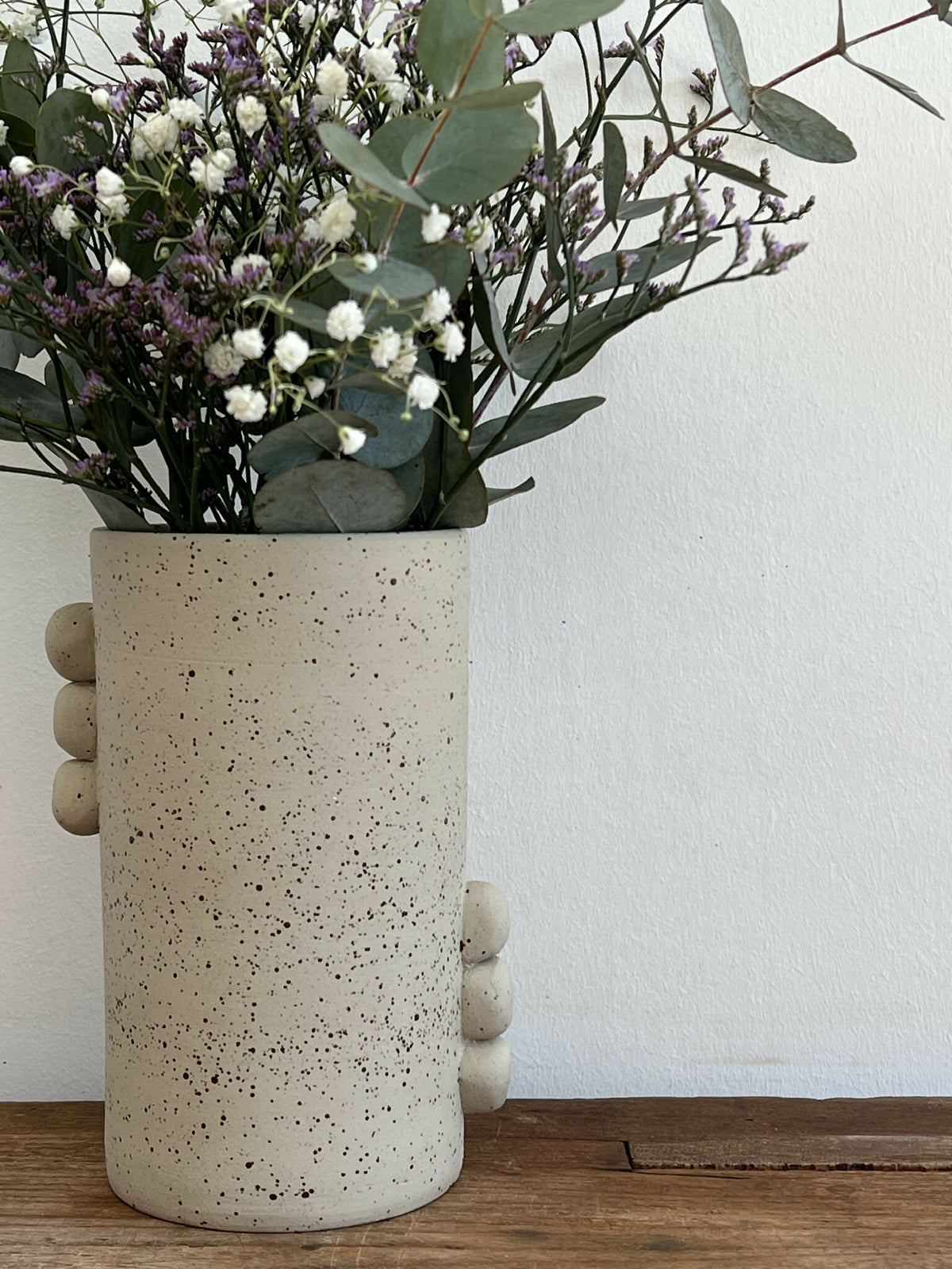 Vase en grès beige piqueté avec bouquet de fleurs blanches et lilas, reliefs ronds.