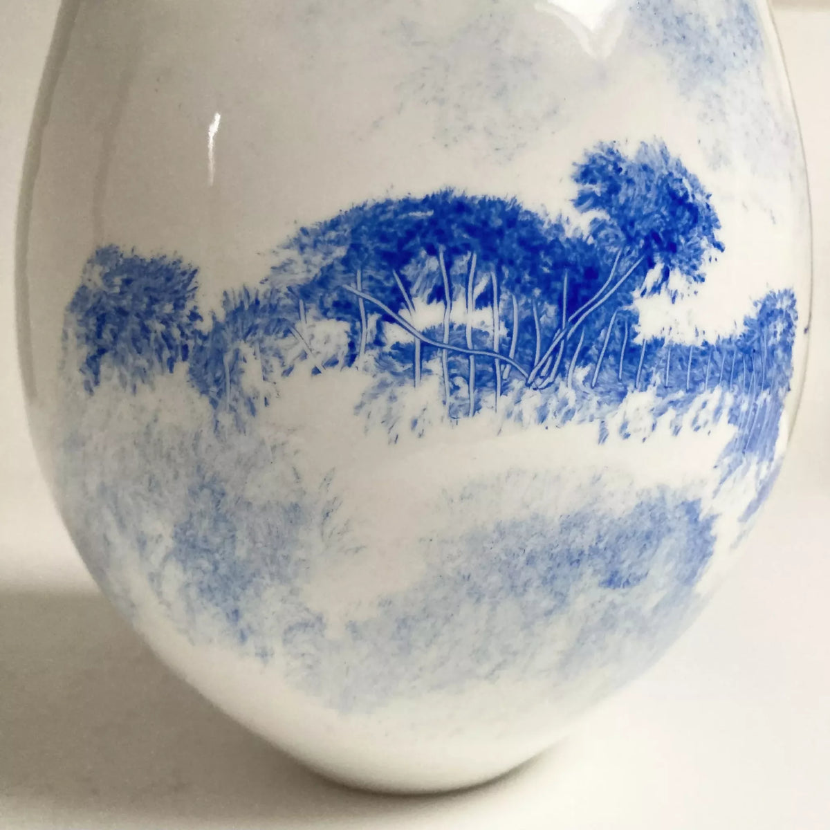 Vase blanc avec motif bleu de paysage et végétation, touches aquarelle.